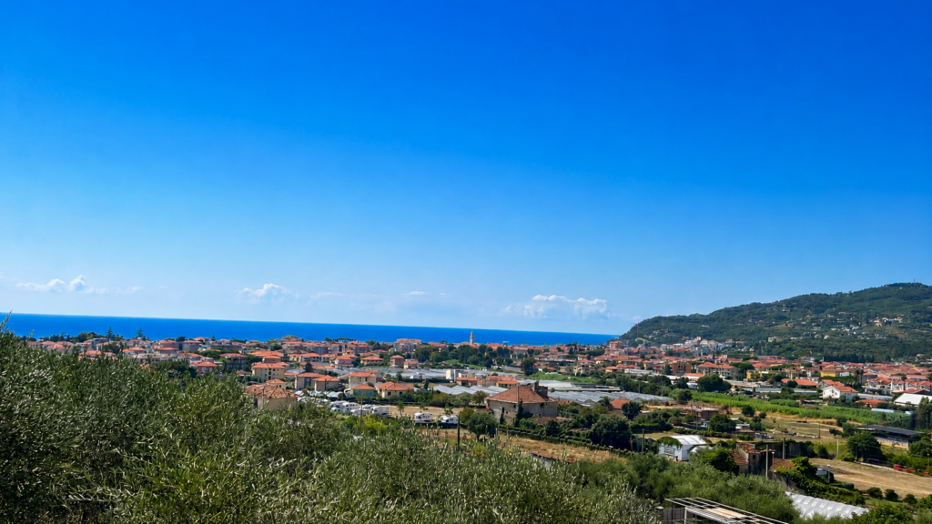 Blick über Olivenhaine auf einen Küstenort in Ligurien mit Meer im Hintergrund